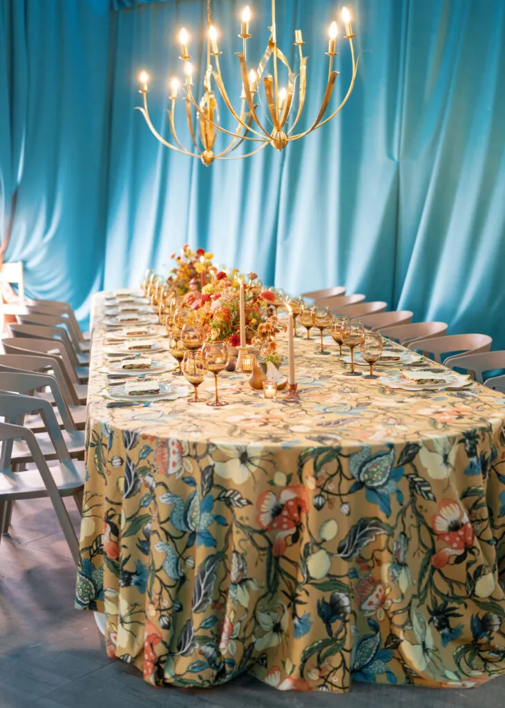 Long table with full floral overlay, amber glassware, colorful centerpieces, and blue draped backdrop. - Curated Linen Collection Elegant wedding table set with floral-patterned linen overlay, surrounded by candles and glassware, under a golden chandelier, against a blue backdrop.