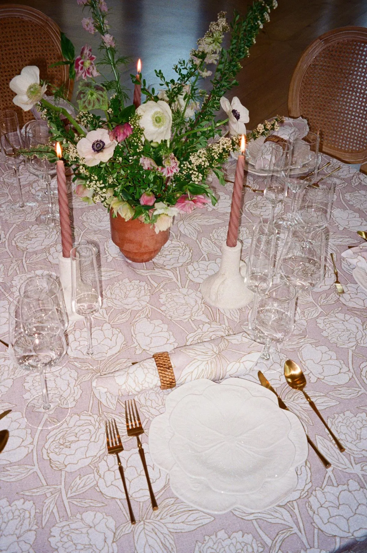 - Curated Linen Collection Elegant wedding tablescape featuring a floral centerpiece with white and pink blooms, soft pastel tablecloth with floral patterns, tapered candles, glassware, and gold cutlery, creating a harmonious and inviting atmosphere.