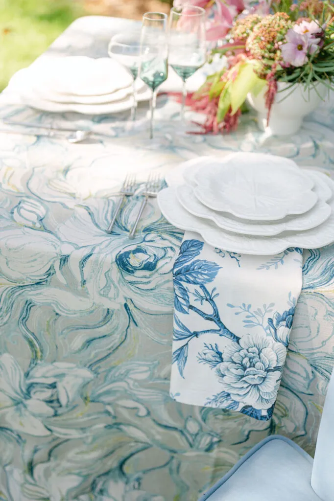 Marbled blue linen with layered white plates and coordinating floral napkin - Curated Linen Collection Elegant table setting featuring a floral patterned linen tablecloth, layered white plates, glassware, and a vibrant floral centerpiece, illustrating texture contrast and color coordination for sophisticated dining experiences.