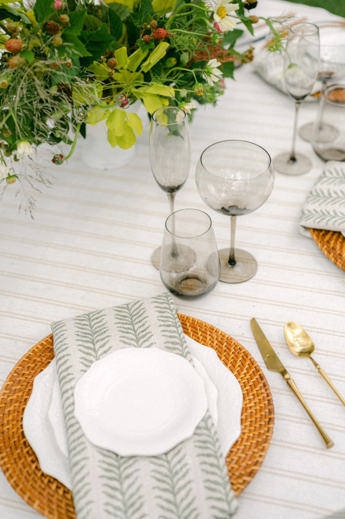 Neutral striped linen table with woven charger, green napkin, and gold flatware - Curated Linen Collection Elegant table setting featuring a layered linen arrangement with a fern-patterned napkin, white plates, woven charger, and assorted glassware, complemented by a vibrant floral centerpiece, showcasing the refined aesthetic of Curated Linen Collection.