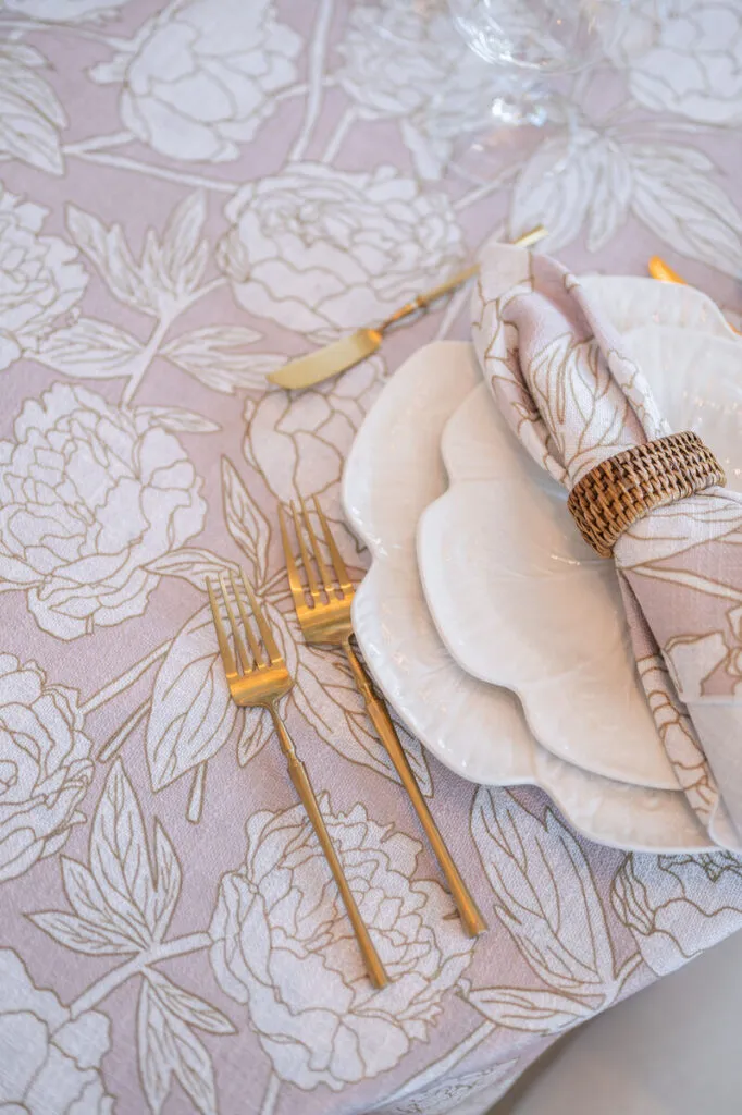 Blush floral linen with gold flatware and layered white plates close-up - Curated Linen Collection Linen table setting featuring a floral patterned tablecloth in soft lavender, elegant white plates, gold flatware, and a neatly arranged napkin with a woven ring, enhancing the sensory and emotional qualities of event design.