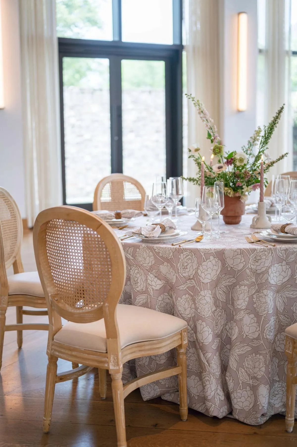 Round table with taupe floral overlay, cane-back chairs, and soft neutral floral centerpiece in bright interior. - Curated Linen Collection Round table with taupe floral overlay, cane-back chairs, and soft neutral floral centerpiece in bright interior.