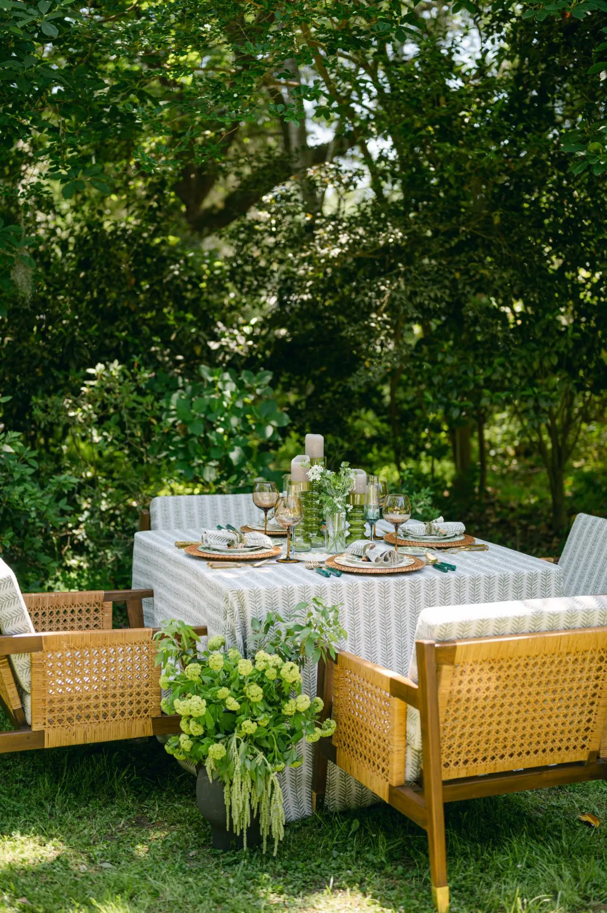 Outdoor table with subtle patterned overlay, cane-back seating, and lush greenery centerpiece.