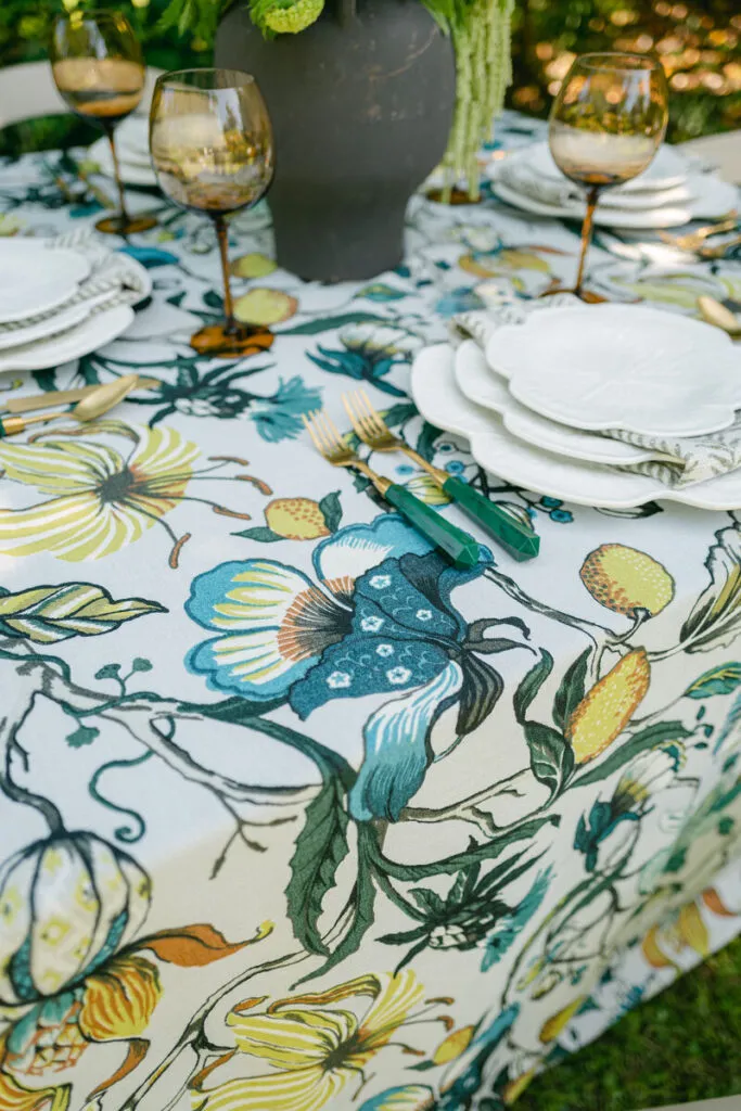Table setting with bold floral linen, white plates, and green-handled flatware - Curated Linen Collection Floral-patterned tablecloth with vibrant colors, elegant dinnerware, and glassware, enhancing a summer tablescape design with breathable fabrics and refined aesthetics.