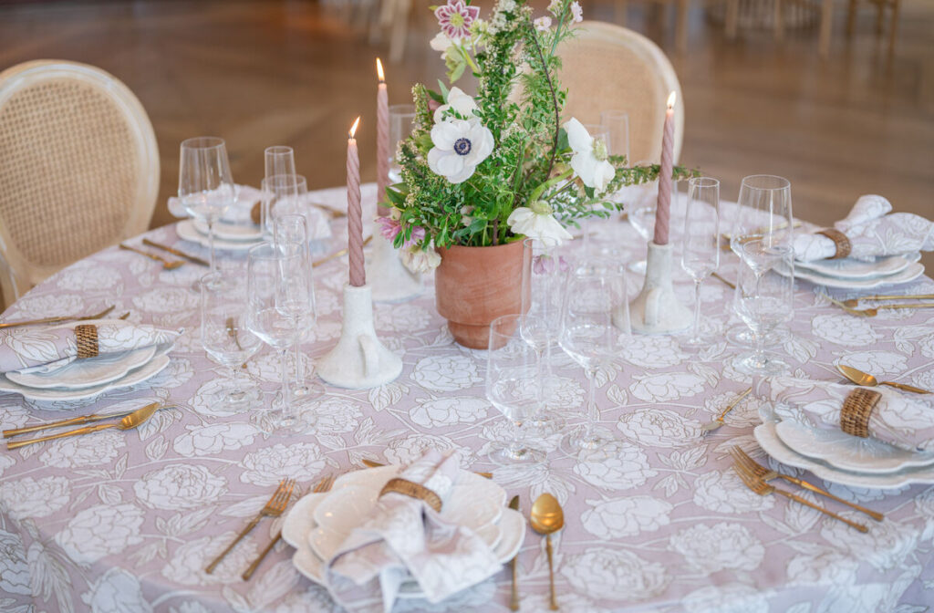 Romantic tablescape with blush floral linen, layered plates, gold flatware, taper candles, and fresh greenery centerpiece. - Curated Linen Collection Romantic tablescape with blush floral linen, layered plates, gold flatware, taper candles, and fresh greenery centerpiece.