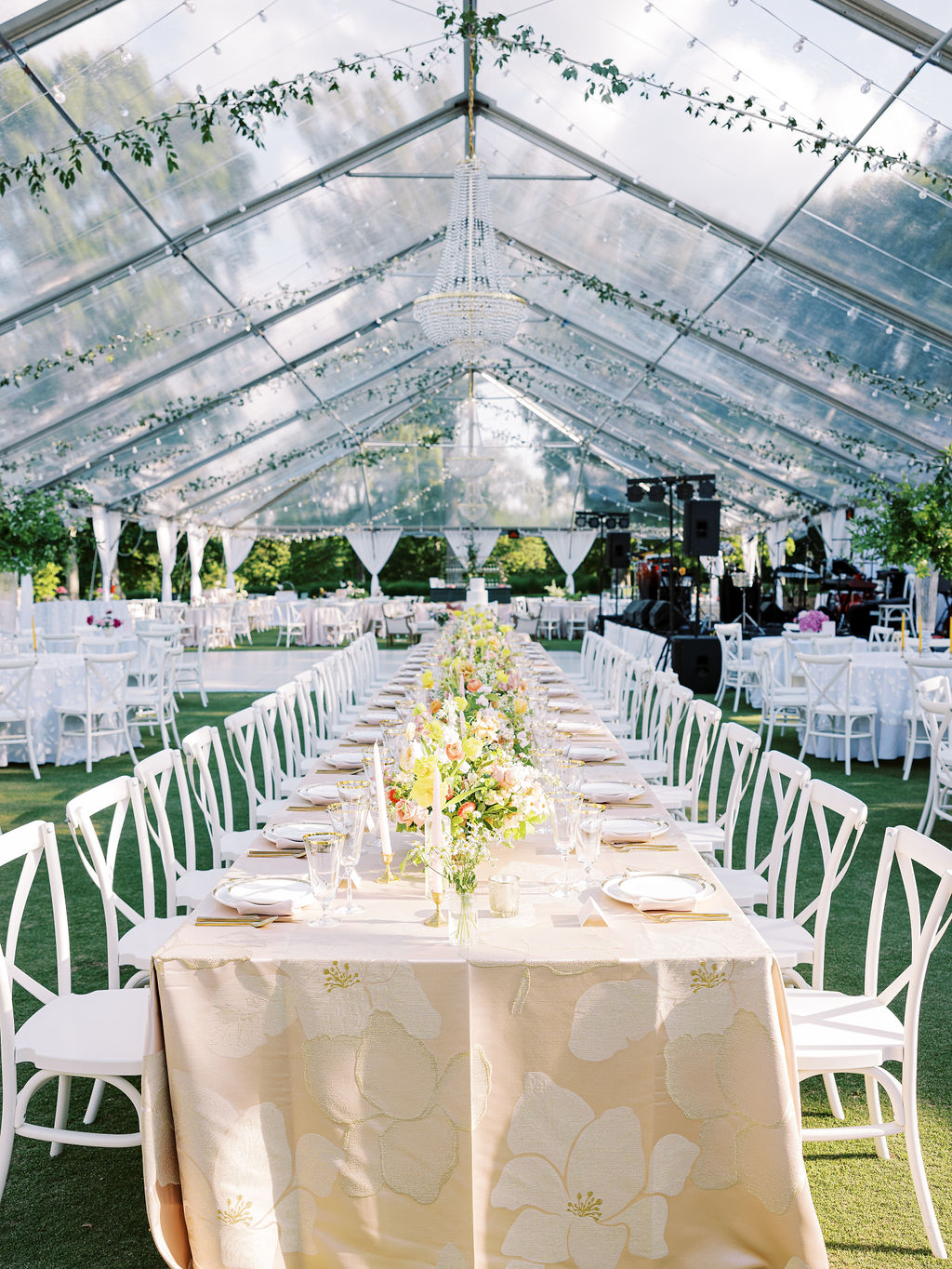 Clear tent wedding reception with long tables, neutral floral linens, white chairs, and hanging greenery. - Curated Linen Collection Clear tent wedding reception with long tables, neutral floral linens, white chairs, and hanging greenery.