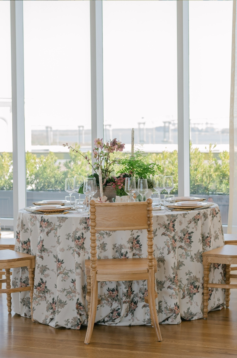 Floral round table linen for a summer wedding reception with wood chairs, glassware, and waterfront views. - Curated Linen Collection Floral patterned linen tablecloth and tableware setting for a wedding reception table.