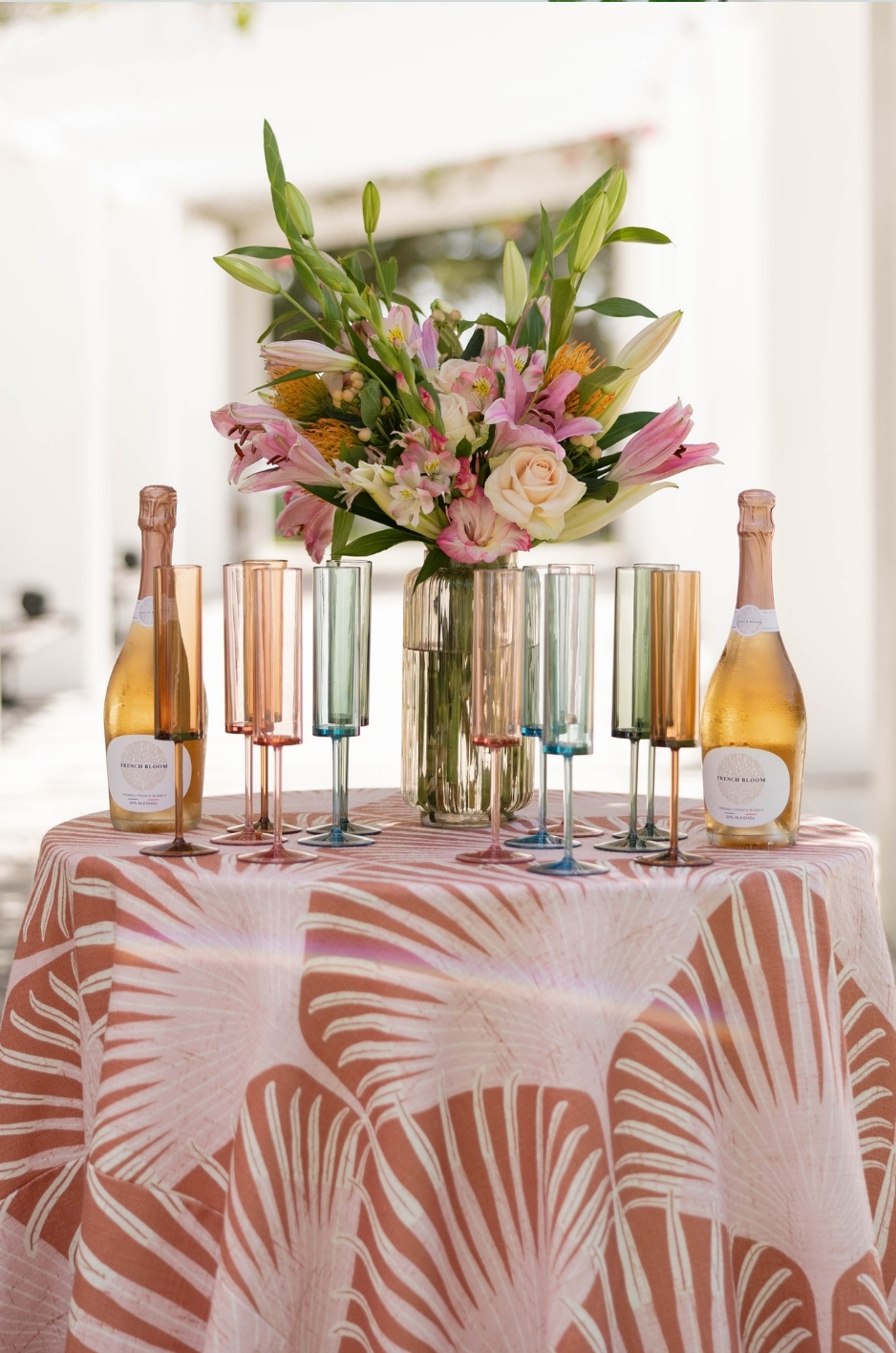 Summer beverage table with coral tropical linen, colorful glassware, champagne bottles, and floral centerpiece. - Curated Linen Collection Summer beverage table with coral tropical linen, colorful glassware, champagne bottles, and floral centerpiece.