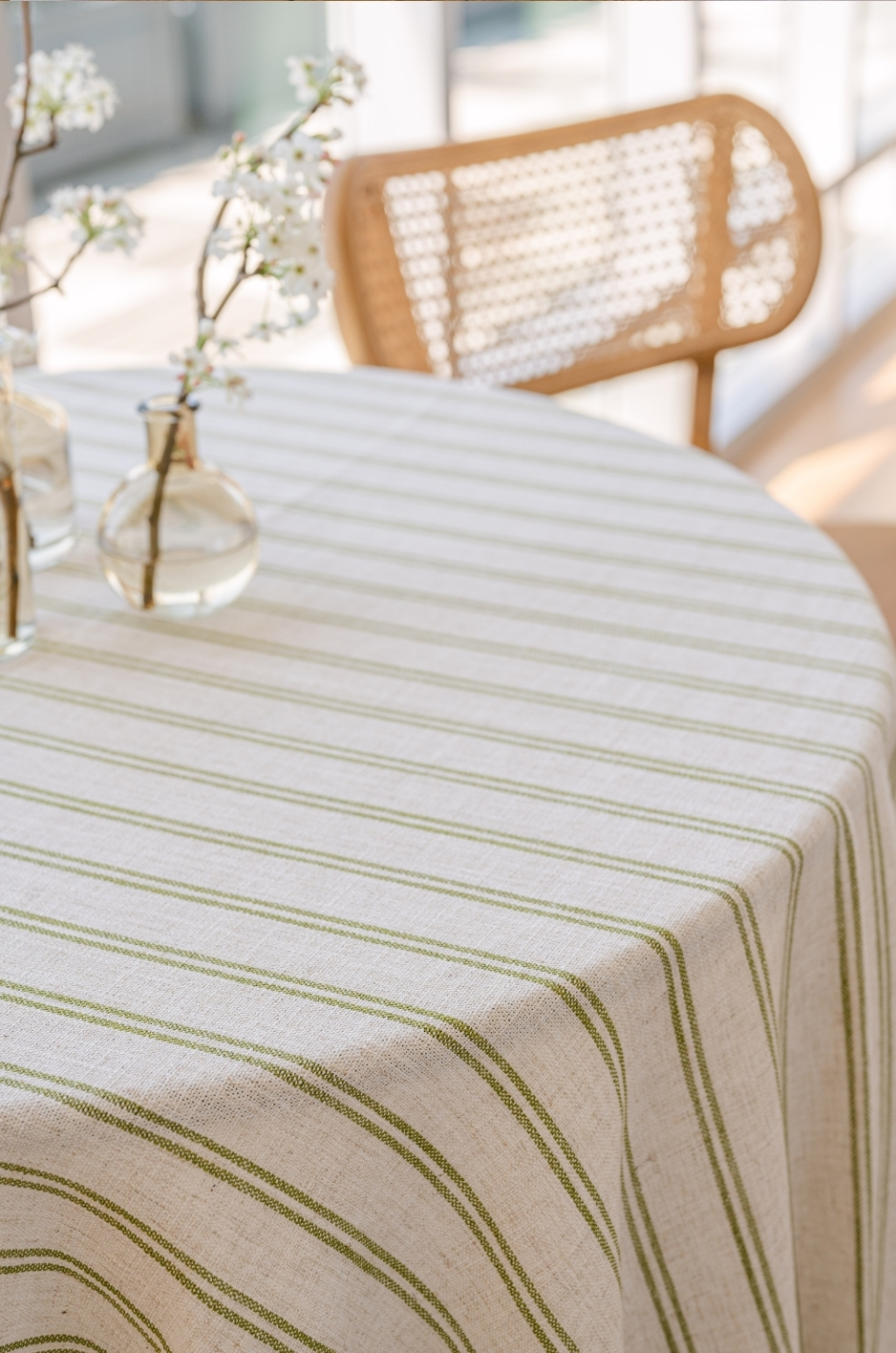 - Curated Linen Collection Sunlit dining table with a beige striped tablecloth, small glass vases of white blossoms, and a wicker chair in the background-to the right.