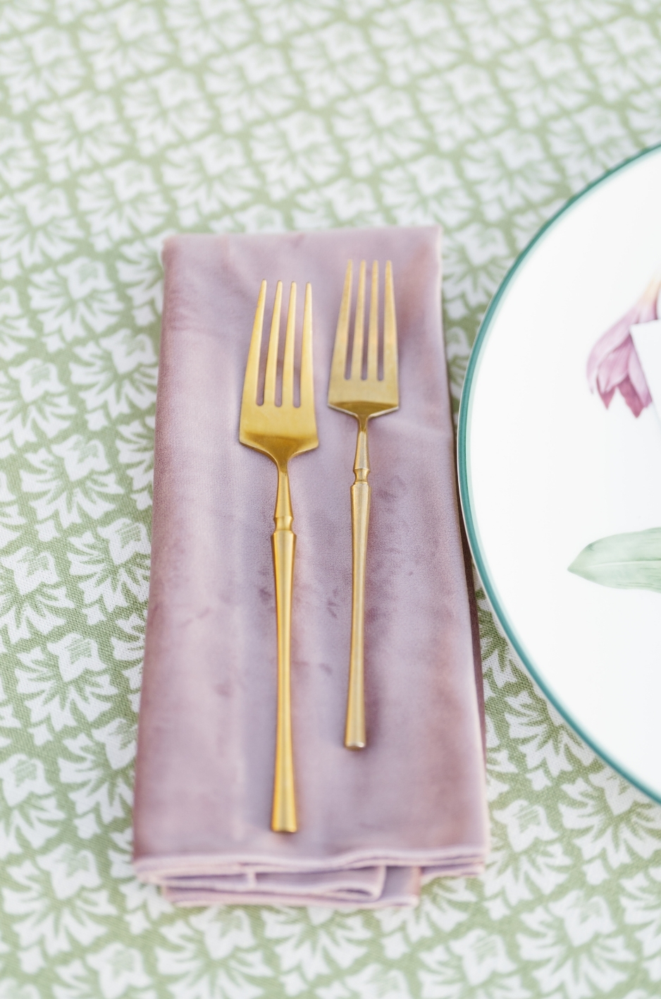 Close-up of gold forks on a mauve napkin over green patterned linen. - Curated Linen Collection Close-up of gold forks on a mauve napkin over green patterned linen.