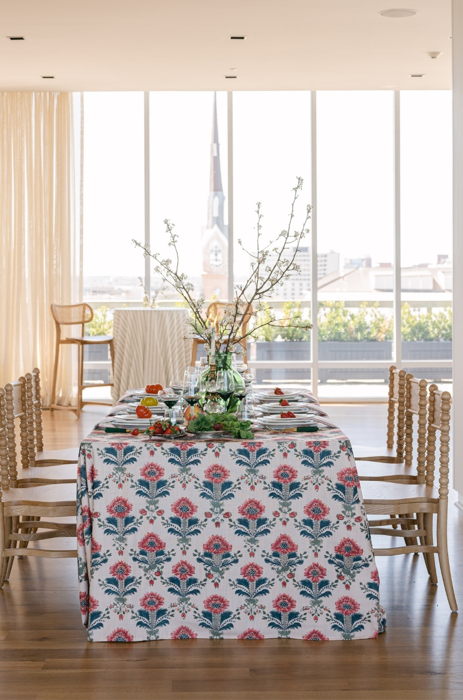 Ballroom tablescape with block-print floral linen, wooden chairs, city views, and tall branch centerpiece. - Curated Linen Collection Ballroom tablescape with block-print floral linen, wooden chairs, city views, and tall branch centerpiece.