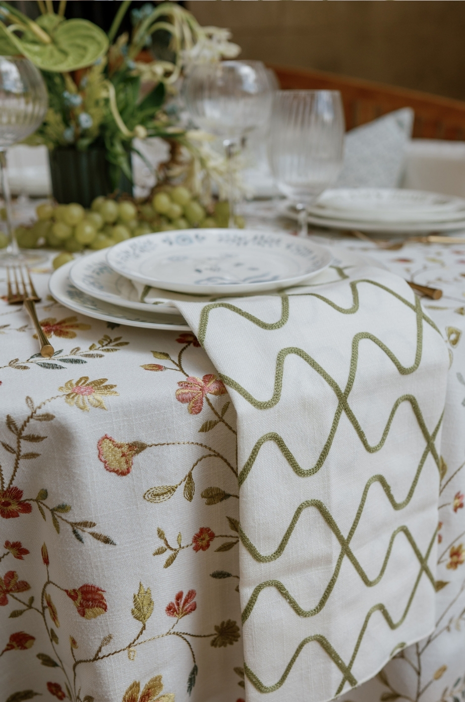 Close-up of floral tablescape with patterned napkin, pale blue plates, glassware, and green grapes. - Curated Linen Collection Close-up of floral tablescape with patterned napkin, pale blue plates, glassware, and green grapes.