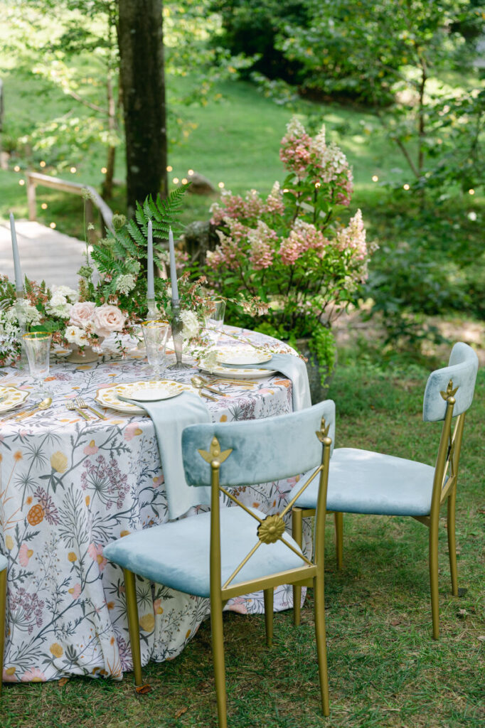 Garden venue tablescape with floral linen, blue velvet chairs, taper candles, and lush summer flowers. - Curated Linen Collection Garden venue tablescape with floral linen, blue velvet chairs, taper candles, and lush summer flowers.