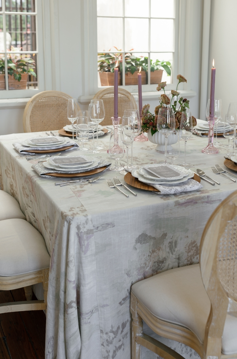 Summer corporate tablescape with soft patterned linen, purple taper candles, layered plates, and woven chairs. - Curated Linen Collection Summer corporate tablescape with soft patterned linen, purple taper candles, layered plates, and woven chairs.