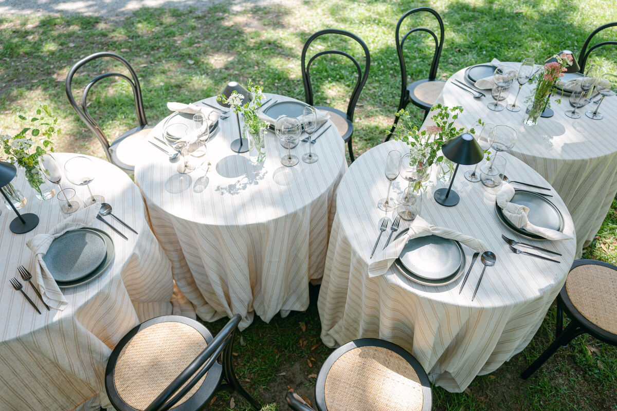Outdoor garden event with round tables dressed in neutral striped linens and black bistro chairs. - Curated Linen Collection Outdoor garden event with round tables dressed in neutral striped linens and black bistro chairs.