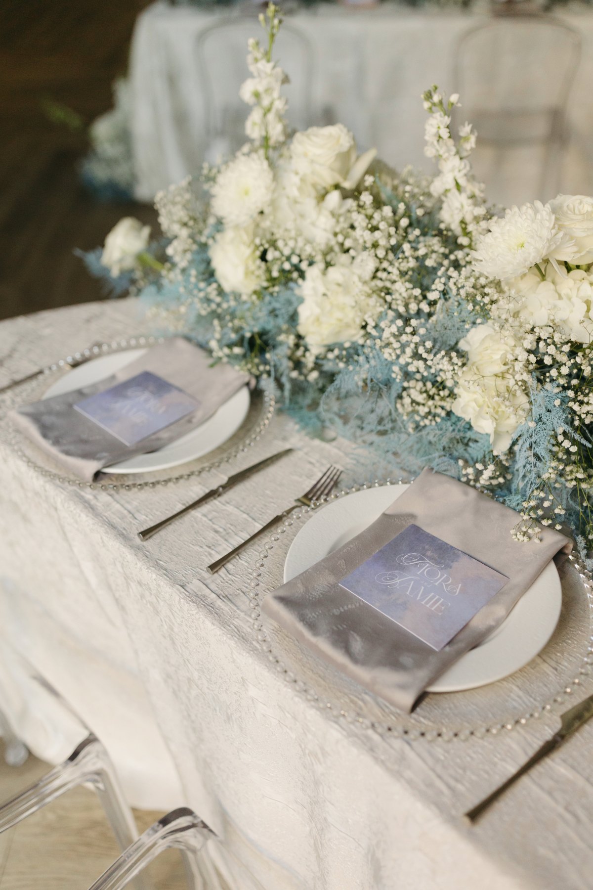 Close-up place settings with white florals, blue accents, silver chargers, and branded menu cards. - Curated Linen Collection Close-up place settings with white florals, blue accents, silver chargers, and branded menu cards.