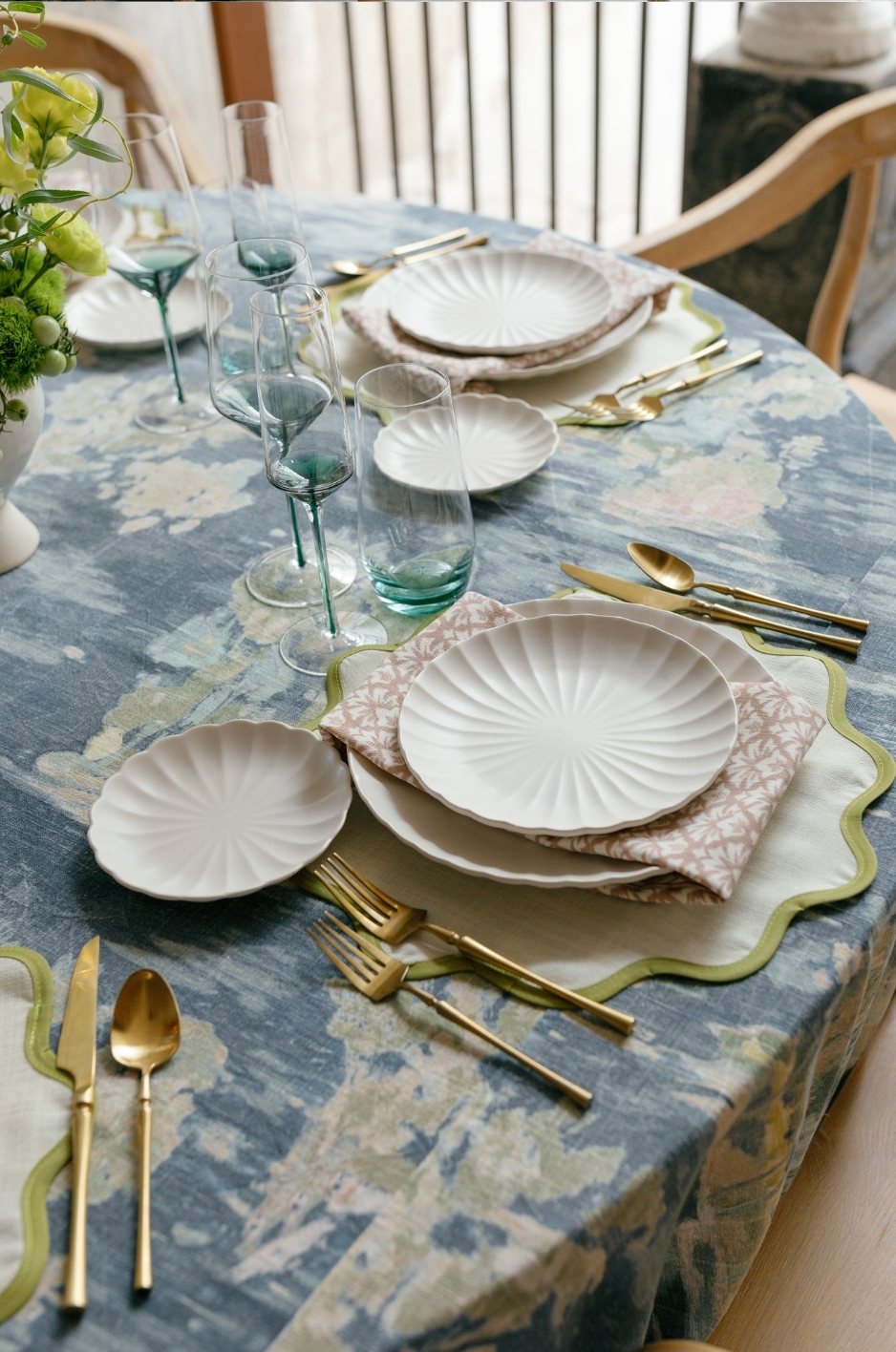 Elegant dining table setting with white plates, gold cutlery, green-tinted glasses, and a pink patterned napkin on a blue floral tablecloth.