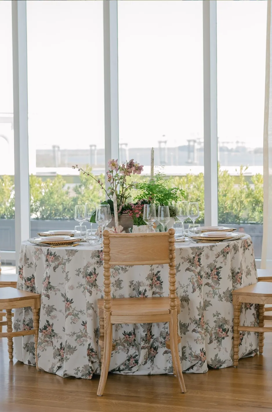 Round banquet table set for a formal event with a floral tablecloth, centerpiece, and glassware in a bright room