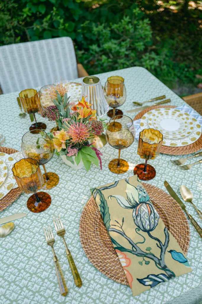 Outdoor event table with textured green linen, amber glassware, woven chargers, and floral napkins - Curated Linen Collection Outdoor event table with textured green linen, amber glassware, woven chargers, and floral napkins
