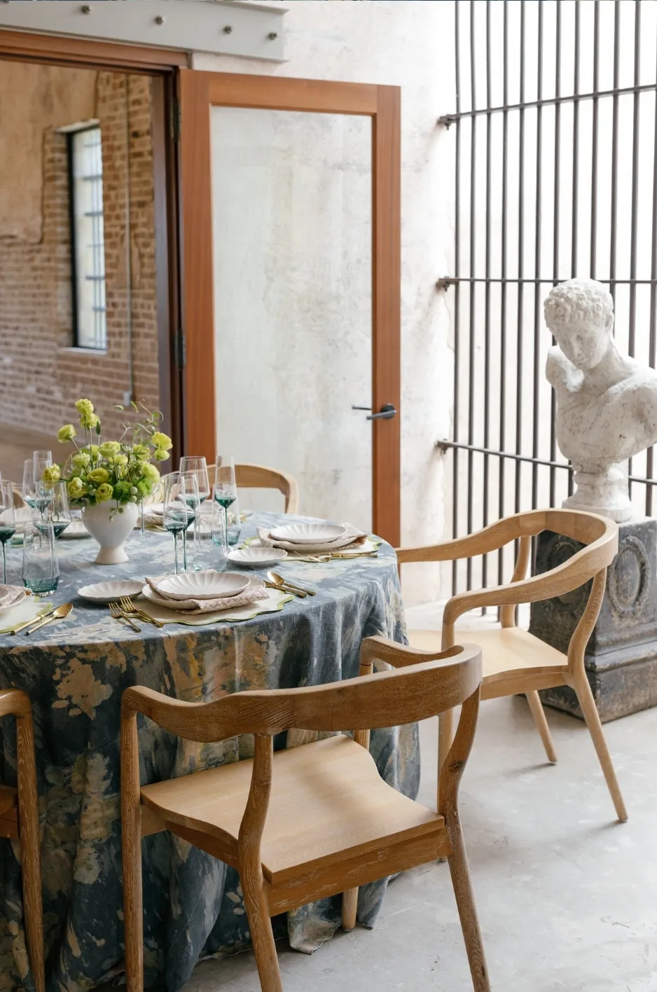 Round wedding table with dark floral linen, wood chairs, layered place settings, and green glassware in an elegant venue - Curated Linen Collection Round wedding table with dark floral linen, wood chairs, layered place settings, and green glassware in an elegant venue