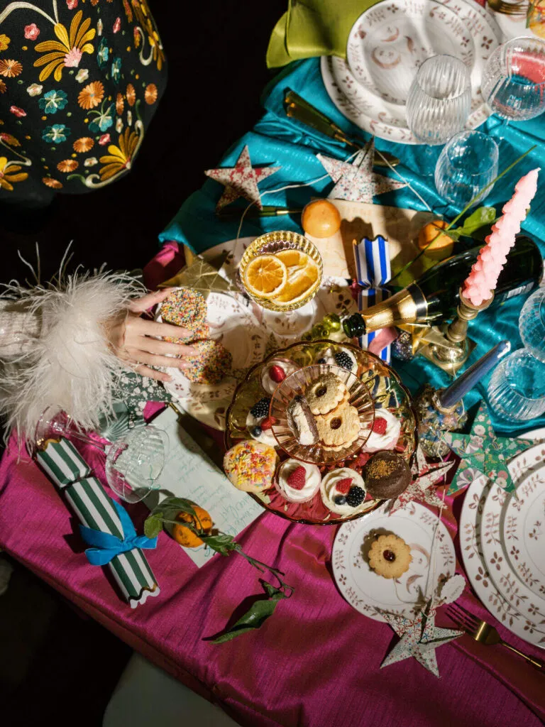 Overhead view of a festive party table with cupcakes, cookies, citrus slices, stars, and a child&rsquo;s hand reaching for treats.