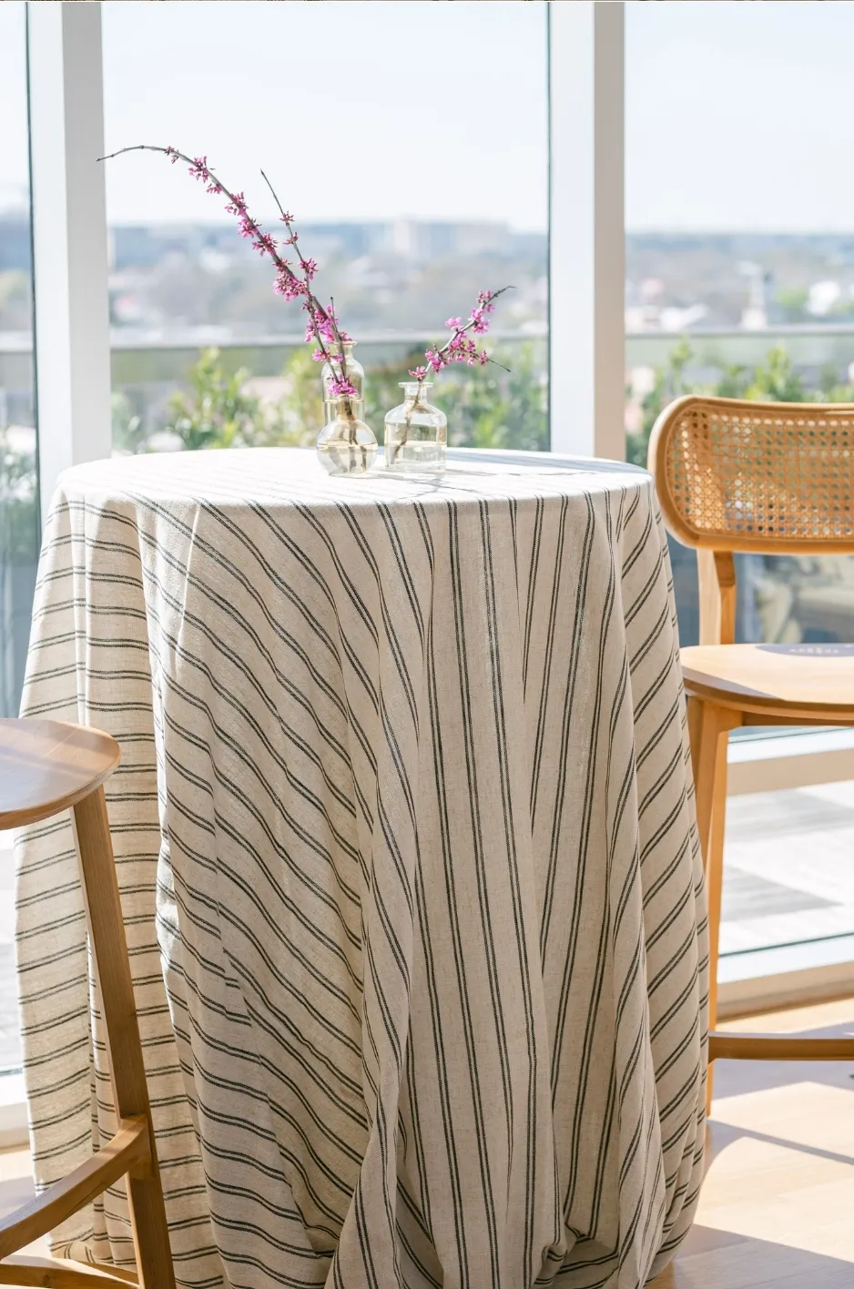 Neutral striped tablecloth on a round wedding table with bud vases and woven chair in a bright venue - Curated Linen Collection Striped linen tablecloth on round table showcasing wedding tablescape design.