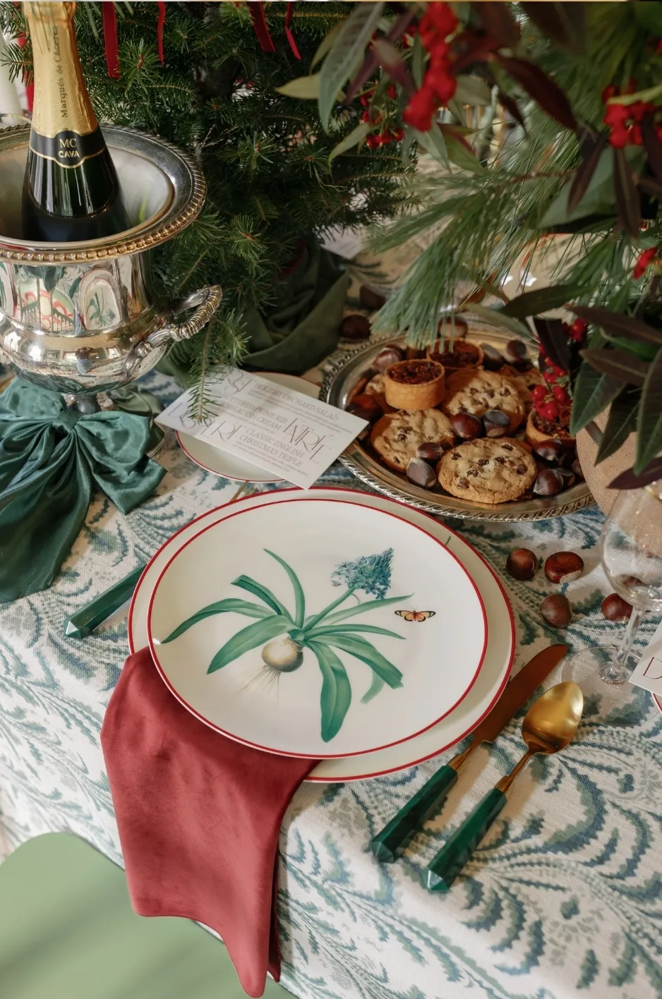 Green patterned tablecloth with botanical china and velvet napkins - Curated Linen Collection Botanical patterned tablecloth and plate setting with red napkin and gold cutlery, avoiding overt seasonal