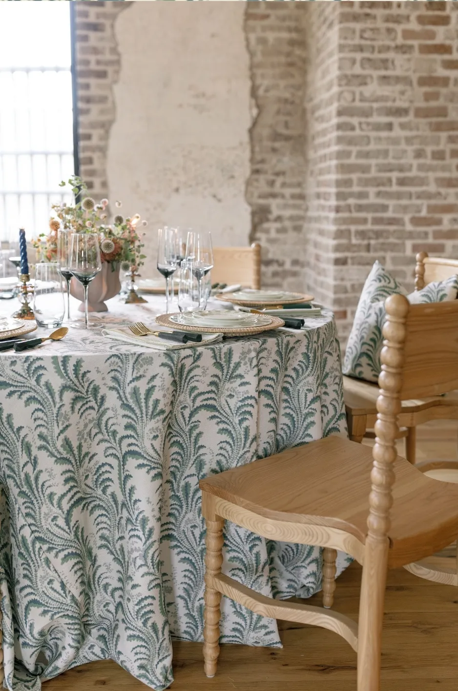 Green patterned wedding tablecloth with wood chairs, layered place settings, glassware, and soft floral centerpiece - Curated Linen Collection Custom wedding tablescape with patterned linen tablecloth, glassware, and floral accents.