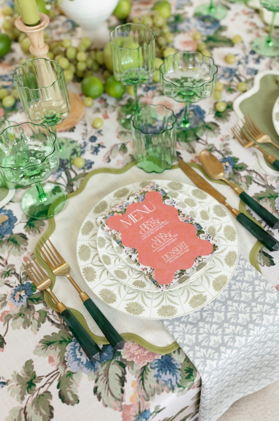 - Curated Linen Collection Green stemware and gold-green flatware frame a floral-patterned plate with a coral menu card on a lively floral tablecloth.
