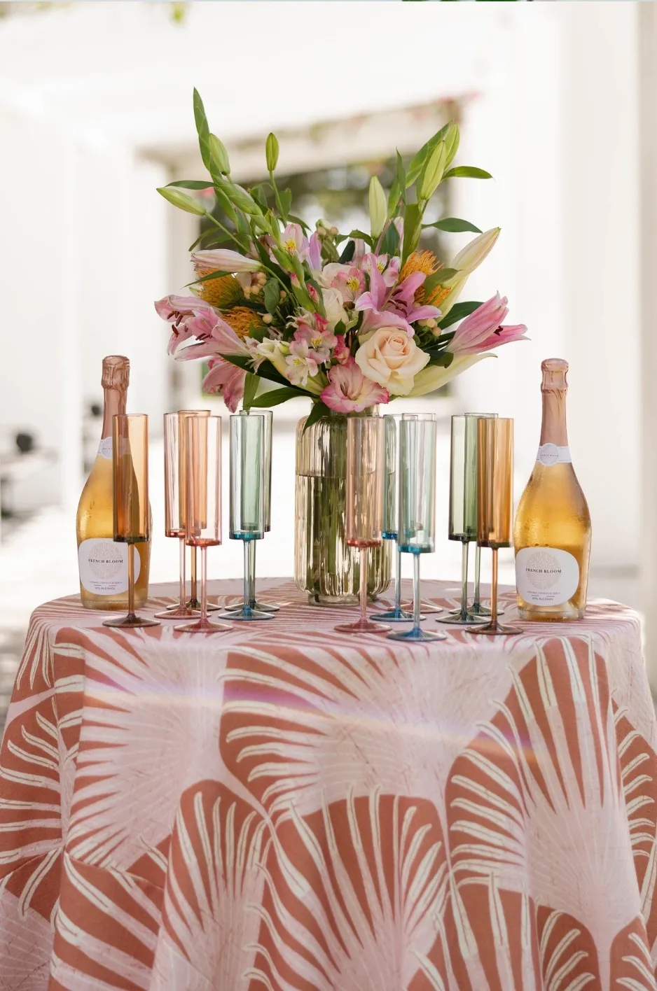 Pink patterned linen table with floral centerpiece, colored glassware, and champagne bottles for a seasonal wedding display - Curated Linen Collection Pink patterned linen table with floral centerpiece, colored glassware, and champagne bottles for a seasonal wedding display