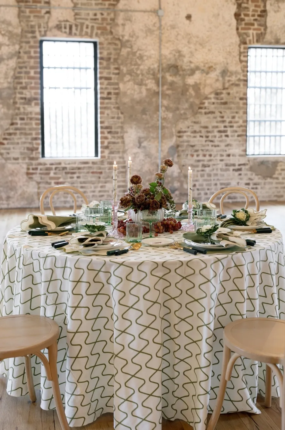 Round reception table with patterned linen, floral centerpiece, and formal place settings in an industrial-style venue - Curated Linen Collection Round reception table with patterned linen, floral centerpiece, and formal place settings in an industrial-style venue