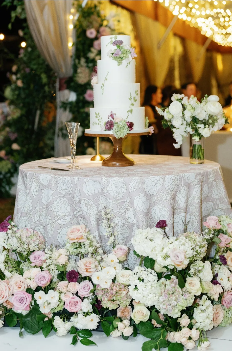 Floral cake table surrounded by white and blush flowers - Curated Linen Collection Luxury wedding cake on linen-draped table with floral arrangements and glassware.