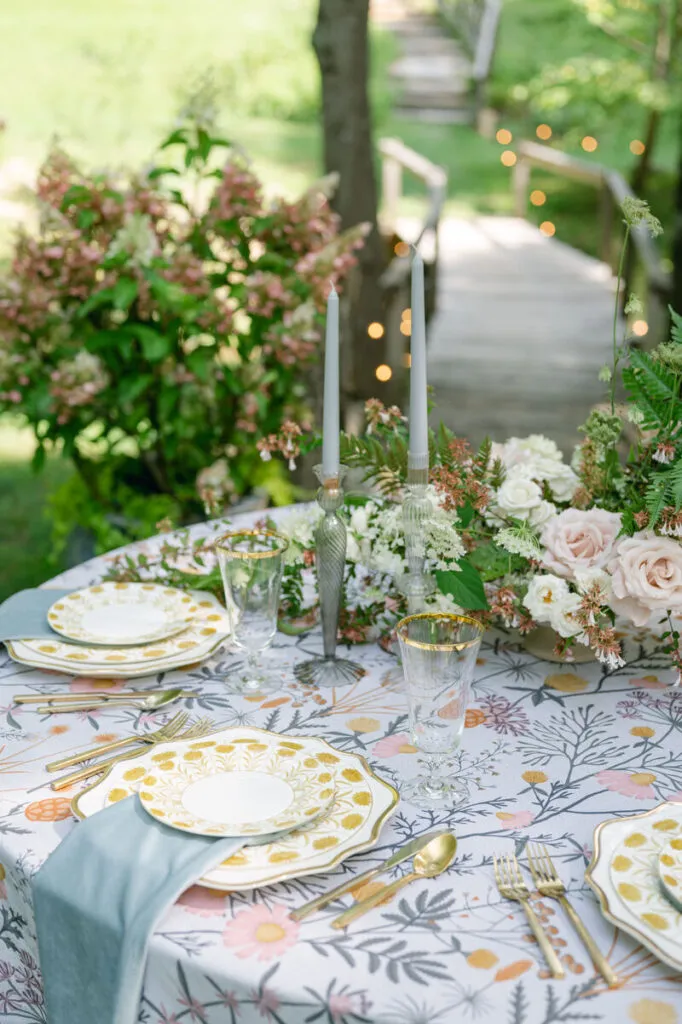 Outdoor wedding table with floral linen, pastel flowers, gold flatware, and garden-style place settings near a wooded bridge - Curated Linen Collection Outdoor wedding table with floral linen, pastel flowers, gold flatware, and garden-style place settings near a wooded bridge