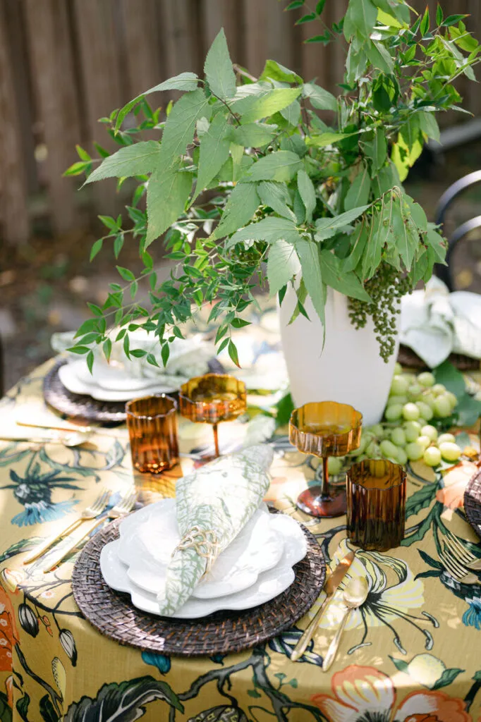Patterned napkin arranged over white plates on a woven charger with amber glassware, gold flatware, and a foliage centerpiece. - Curated Linen Collection Patterned napkin arranged over white plates on a woven charger with amber glassware, gold flatware, and a foliage centerpiece.
