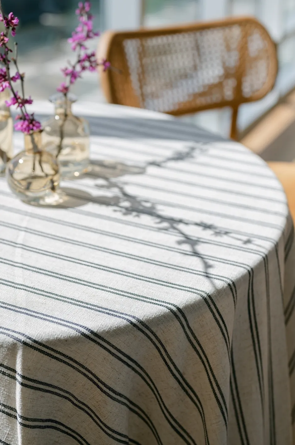 - Curated Linen Collection Round table with a striped linen tablecloth, small glass vases holding pink blossoms, and a wicker chair in a sunlit outdoor setting.
