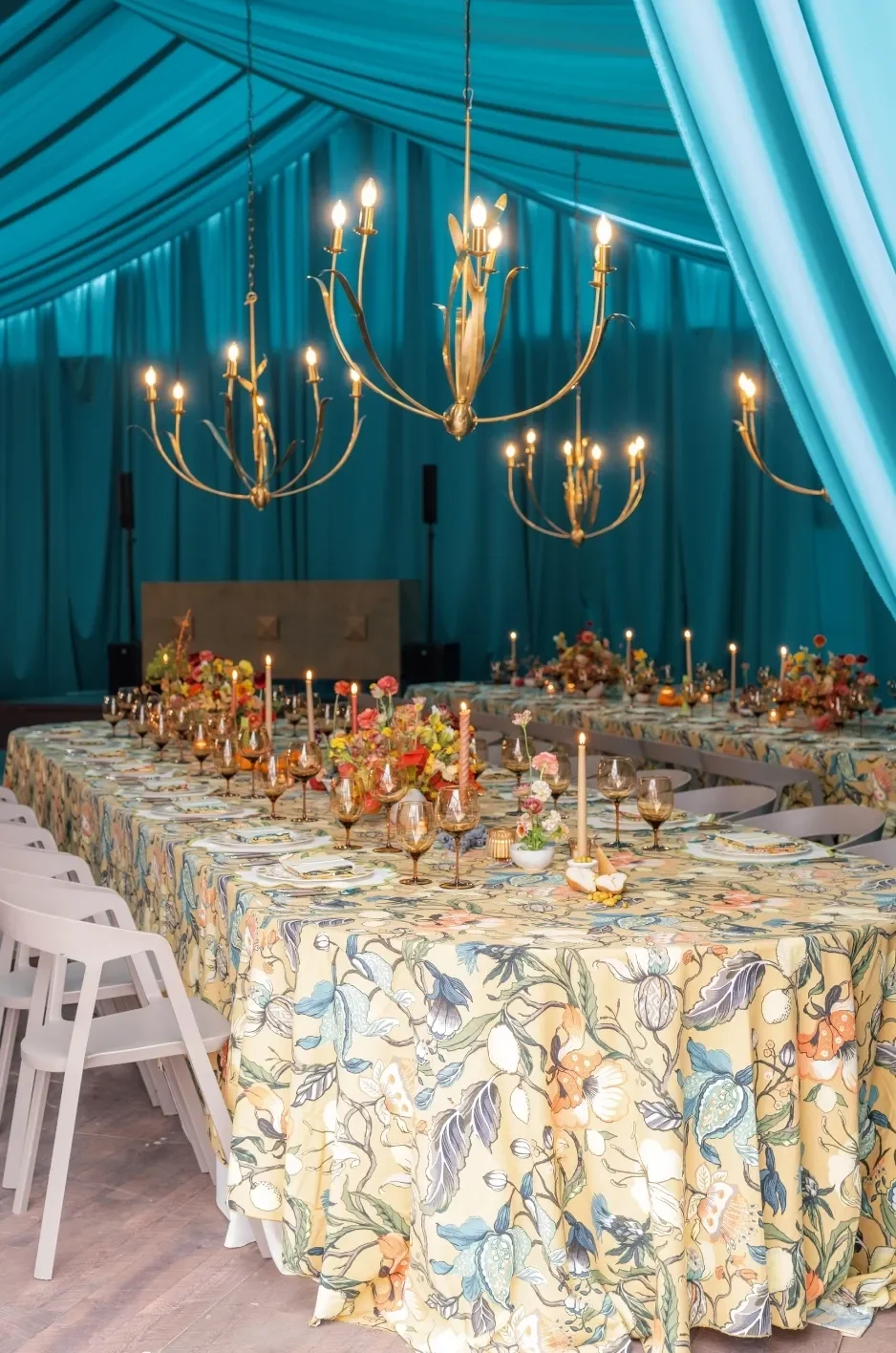 U-shaped reception tables covered in botanical linens beneath teal draping, gold chandeliers, and candlelight. U-shaped reception tables covered in botanical linens beneath teal draping, gold chandeliers, and candlelight.
