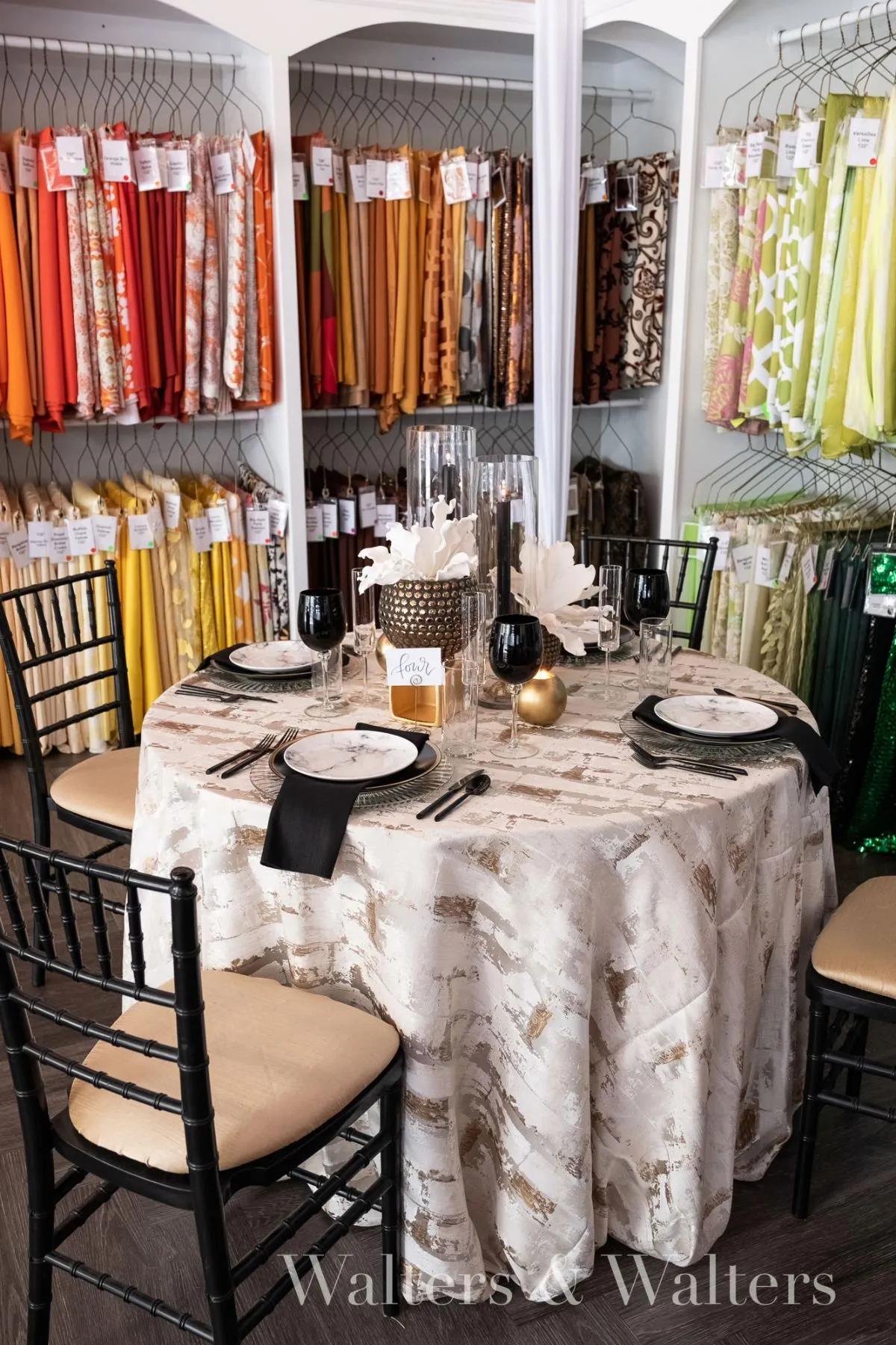 Linen showroom table with neutral patterned cloth, black napkins, black goblets, white place settings, and colorful fabric samples displayed behind it. - Curated Linen Collection Linen showroom table with neutral patterned cloth, black napkins, black goblets, white place settings, and colorful fabric samples displayed behind it.