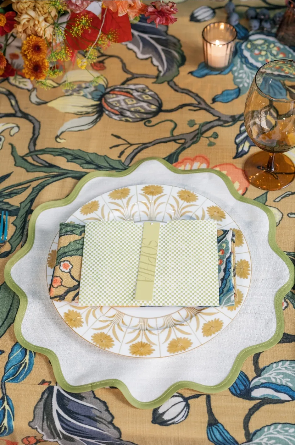 Close-up place setting on bold botanical linen with scalloped charger, patterned china, menu, florals, and amber glassware