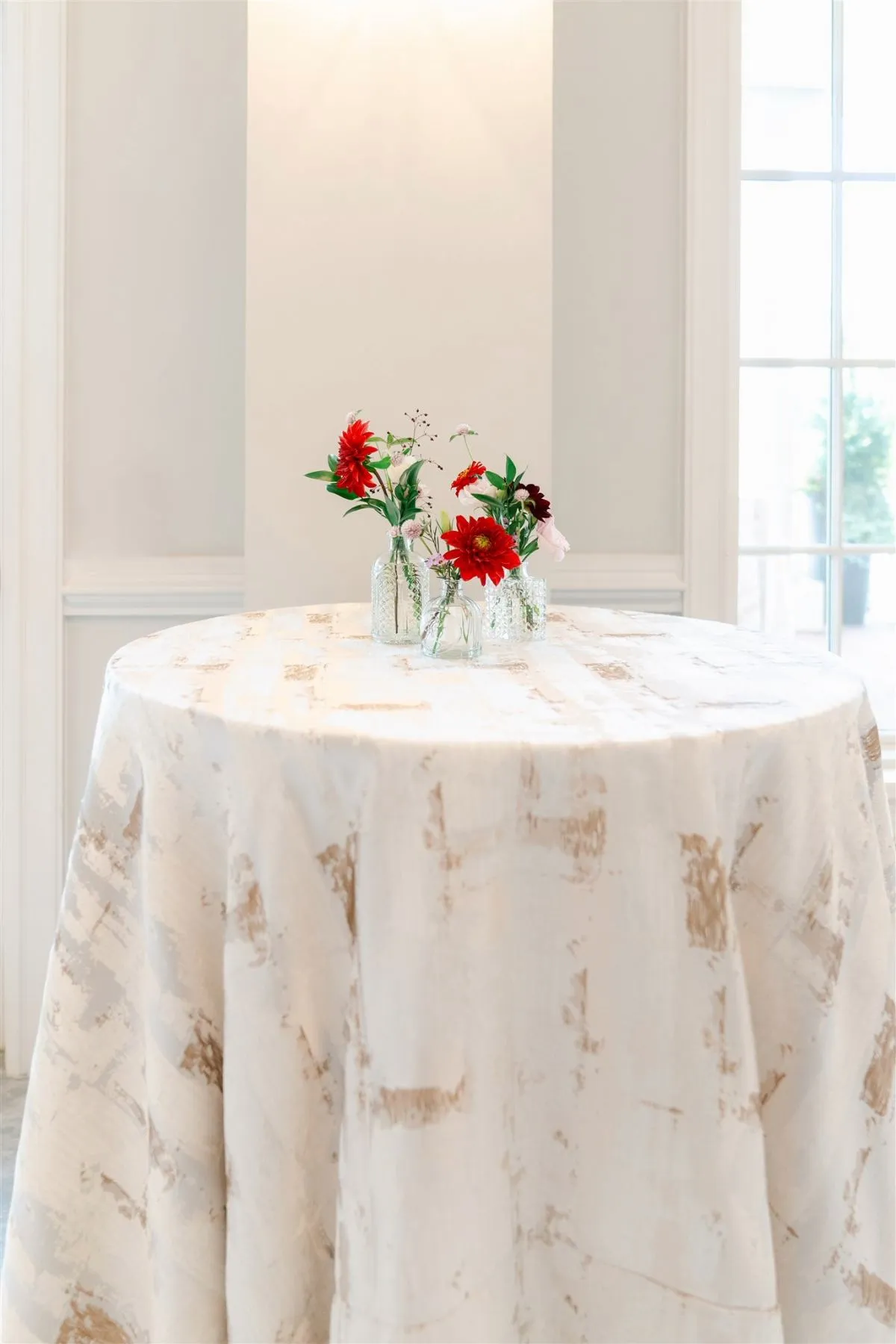 - Curated Linen Collection Round table with a white/cream marbled tablecloth and glass vases holding red and pink flowers as a centerpiece in a bright room.
