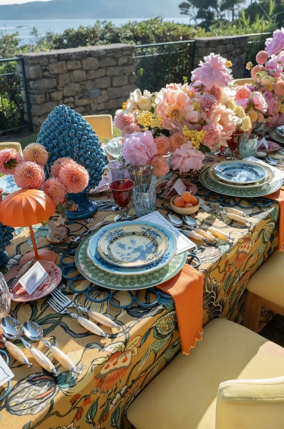 Outdoor reception table with an ornate multicolor linen, orange napkins, blue china, and abundant pink florals. Outdoor reception table with an ornate multicolor linen, orange napkins, blue china, and abundant pink florals.