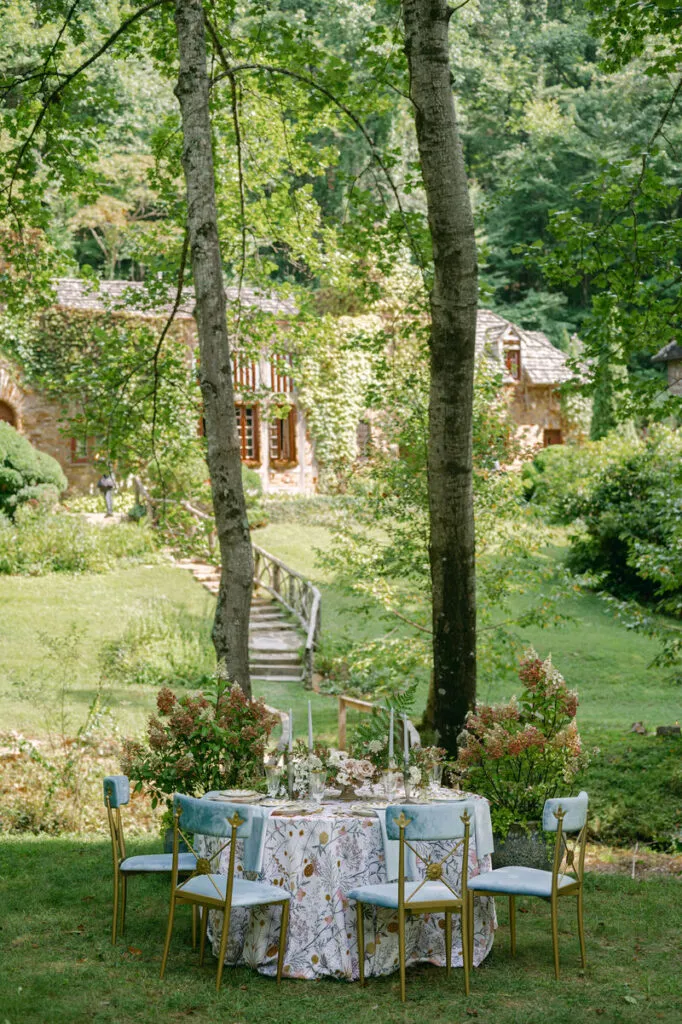 Garden dining table with a floral linen, gold-framed chairs, candles, and seasonal flowers overlooking a wooded estate. - Curated Linen Collection Garden dining table with a floral linen, gold-framed chairs, candles, and seasonal flowers overlooking a wooded estate.