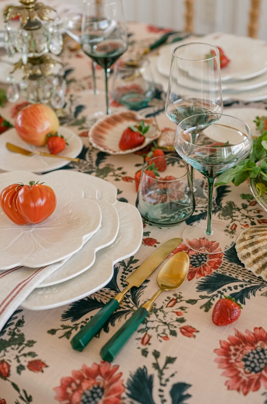 Floral table linen with layered white plates, green-handled flatware, blue-green glassware, tomatoes, strawberries, and shell-shaped dishes. - Curated Linen Collection Floral table linen with layered white plates, green-handled flatware, blue-green glassware, tomatoes, strawberries, and shell-shaped dishes.