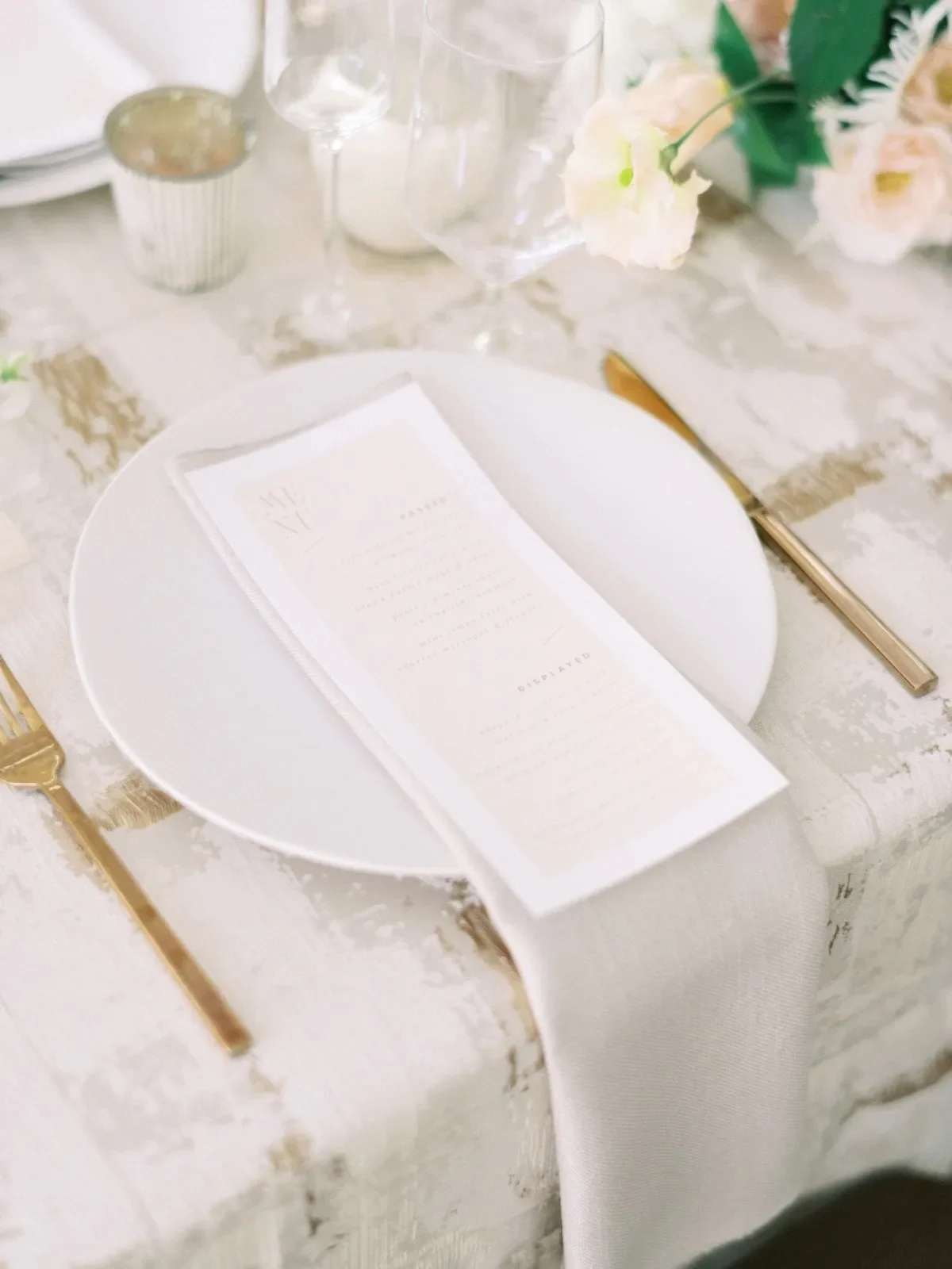 Neutral wedding napkin folded beneath a menu card on white dinnerware with gold flatware and pale florals. Neutral wedding napkin folded beneath a menu card on white dinnerware with gold flatware and pale florals.