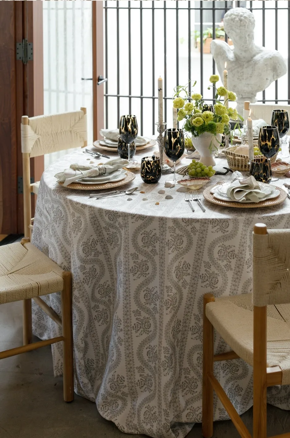 Round reception table with patterned linen, black goblets, white napkins, woven chairs, green flowers, and a classical bust. - Curated Linen Collection Round reception table with patterned linen, black goblets, white napkins, woven chairs, green flowers, and a classical bust.