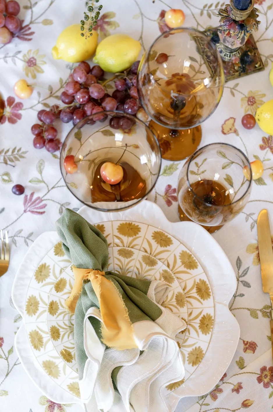 Green and cream napkin gathered with a mustard ribbon over floral china beside amber goblets, grapes, and lemons. - Curated Linen Collection Green and cream napkin gathered with a mustard ribbon over floral china beside amber goblets, grapes, and lemons.