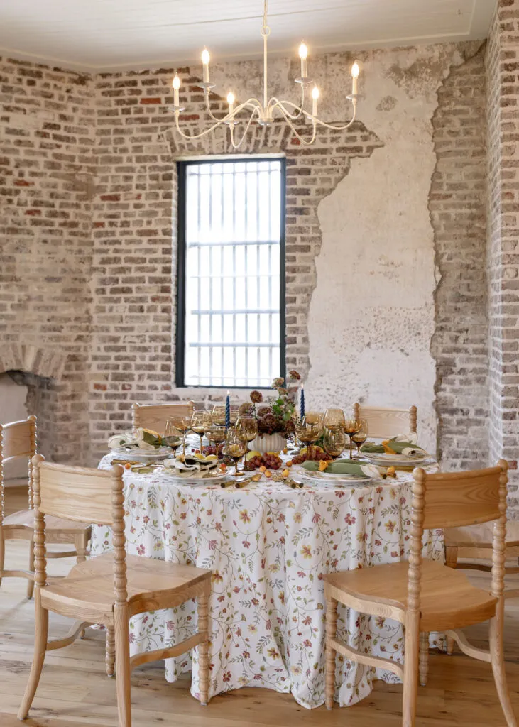 Round dining table with a delicate floral linen, light wood chairs, amber glassware, seasonal fruit, and a chandelier in a historic brick room.