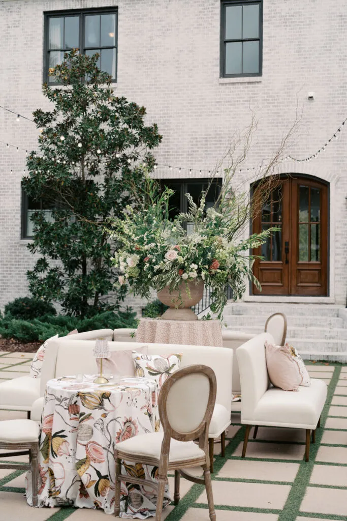 Outdoor courtyard lounge with a botanical table linen, upholstered seating, a large floral arrangement, string lights, and a brick building. - Curated Linen Collection Outdoor courtyard lounge with a botanical table linen, upholstered seating, a large floral arrangement, string lights, and a brick building.