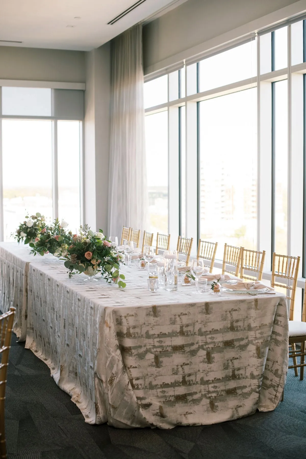 Long rectangular banquet table set for a formal event with white floral centerpieces and gold chairs by large windows in a bright room