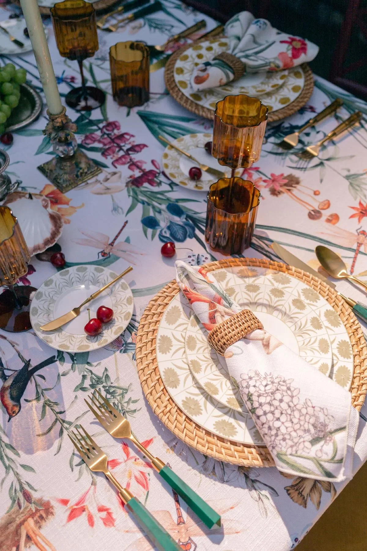 Botanical printed napkin secured with a woven ring beside patterned china, amber glassware, and green flatware. Botanical printed napkin secured with a woven ring beside patterned china, amber glassware, and green flatware.