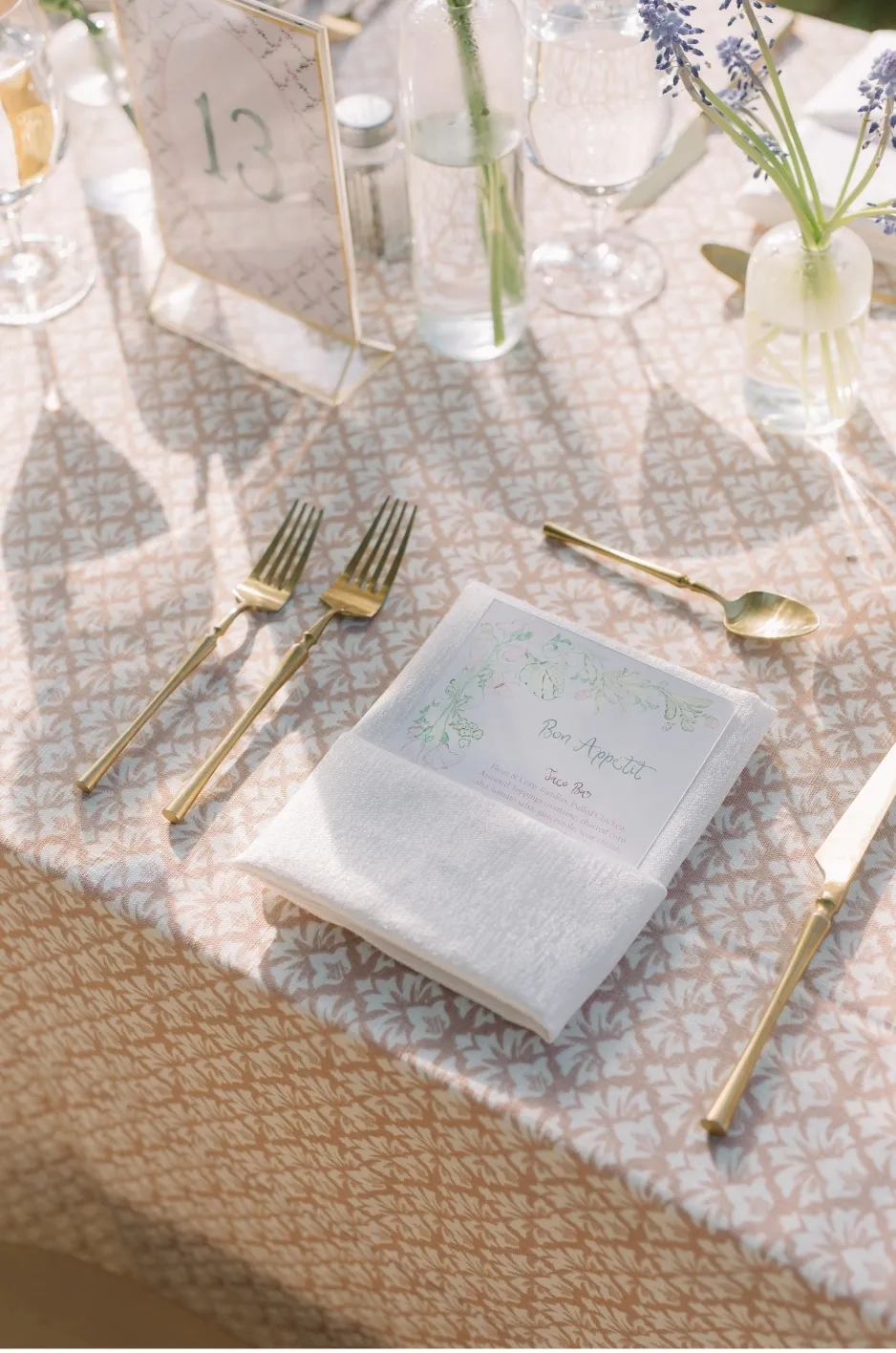 Elegant table setting with gold forks and spoon, white napkin, and a menu card on a beige patterned tablecloth, lavender bouquets in glass vases.